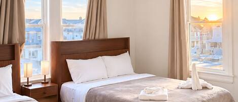 A hotel room with two beds, a wooden headboard, a nightstand with a lamp, and a view of the city through large windows.