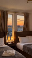 Habitación de hotel con dos camas, una ventana con cortinas, una mesita de noche con lámparas y vistas a edificios.