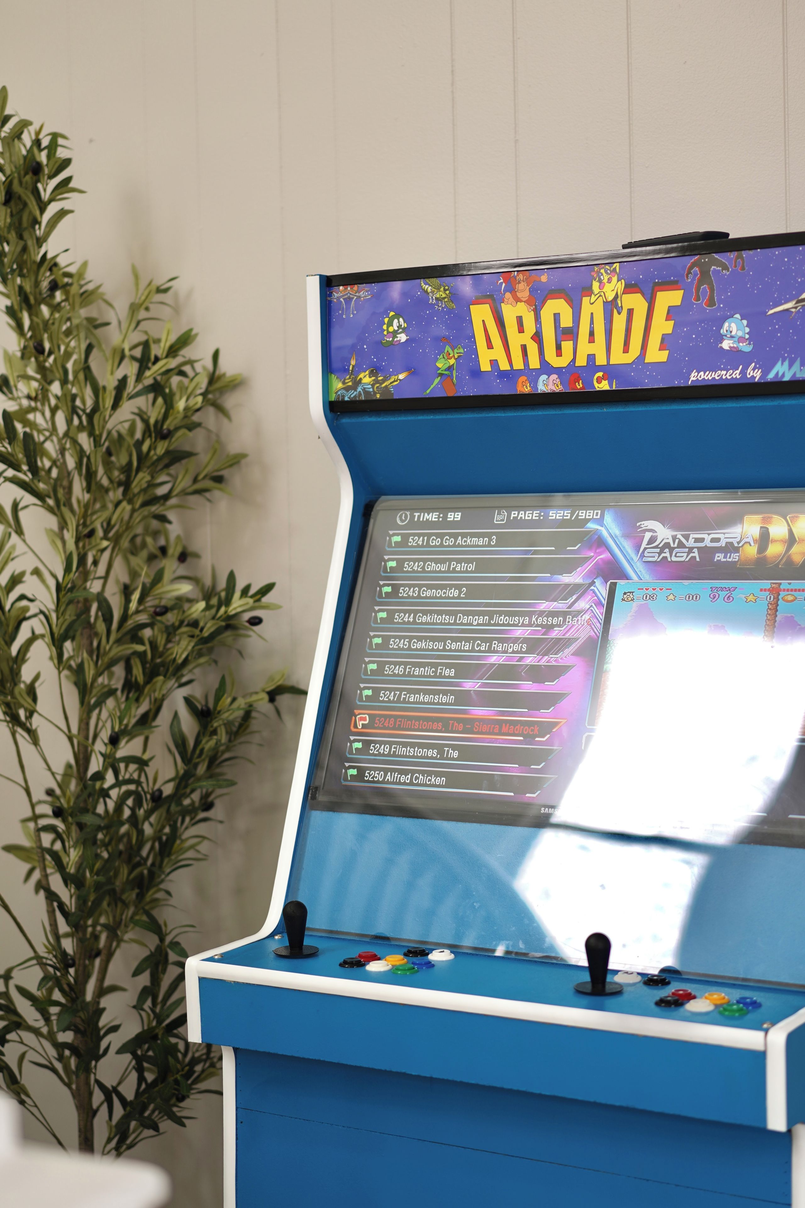 A blue arcade machine with a screen displaying a game menu, surrounded by a plant.
