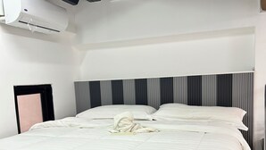 A hotel room with a large bed, a headboard with vertical stripes, an air conditioning unit on the wall, and a window with blinds.