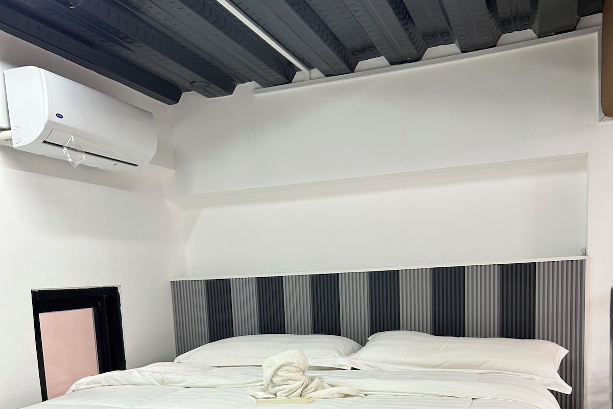 Una habitación de hotel con una cama grande, un cabecero con rayas verticales, un equipo de aire acondicionado en la pared y una ventana con persianas.