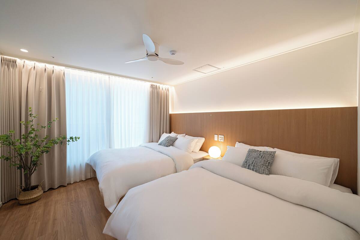 A hotel room with two beds, a ceiling fan, a bedside lamp, and a potted plant.
