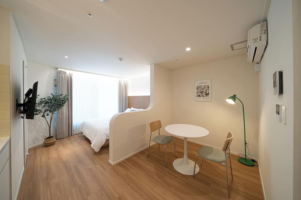 A modern hotel room with a bed, a dining table with chairs, a potted plant, and a wall-mounted air conditioner.