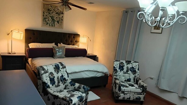 Un dormitorio con una cama, dos sillas, una mesita de noche, un ventilador de techo y una lámpara de araña.