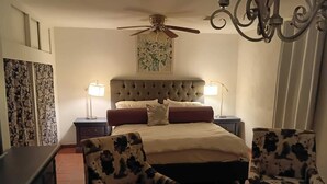Un dormitorio con un cabecero capitonado, dos mesitas de noche con lámparas, una cama con colcha color beige, dos sillones con estampado floral, un ventilador de techo y una araña.