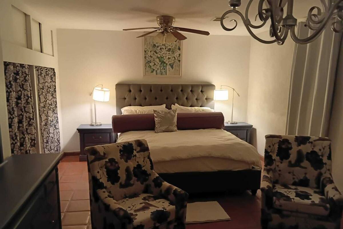 Une chambre à coucher avec une tête de lit capitonnée, deux tables de chevet avec des lampes, un lit recouvert d’une couvre-lit beige, deux fauteuils à motifs floraux, un ventilateur de plafond et un lustre.