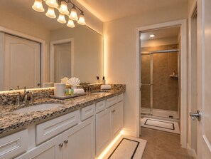 A bathroom with a granite countertop, white cabinetry, a large mirror, and a shower with beige tiles.
