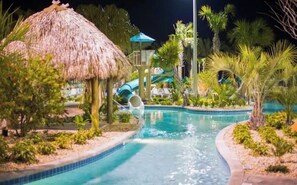 A swimming pool with a water slide, surrounded by tropical vegetation and a thatched-roof structure.