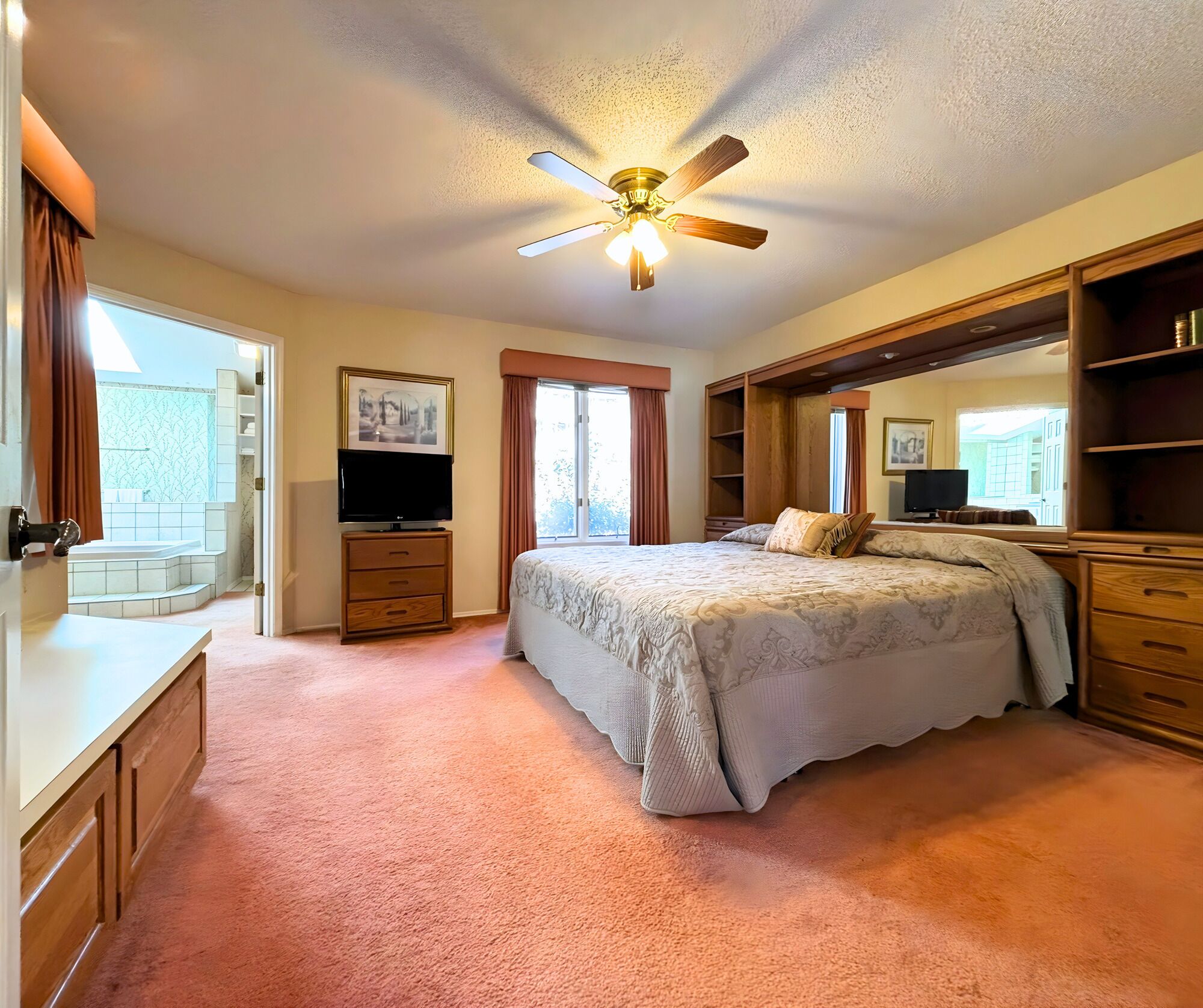 A bedroom with a bed, a ceiling fan, a TV, a dresser, and a large window.