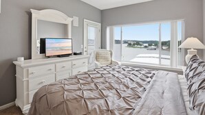 Une chambre avec un grand lit, un téléviseur posé sur une commode, une chaise, une fenêtre donnant sur un plan d’eau et une lampe.