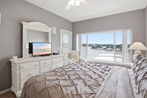 Ein Schlafzimmer mit einem großen Bett, einem Fernseher auf einer Kommode, einem Sessel, einem Fenster mit Blick auf ein Gewässer und einer Lampe.
