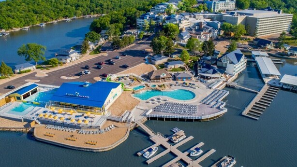 Vue aérienne d’un complexe hôtelier situé au bord de l’eau, avec une grande piscine, plusieurs bâtiments et des bateaux amarrés.