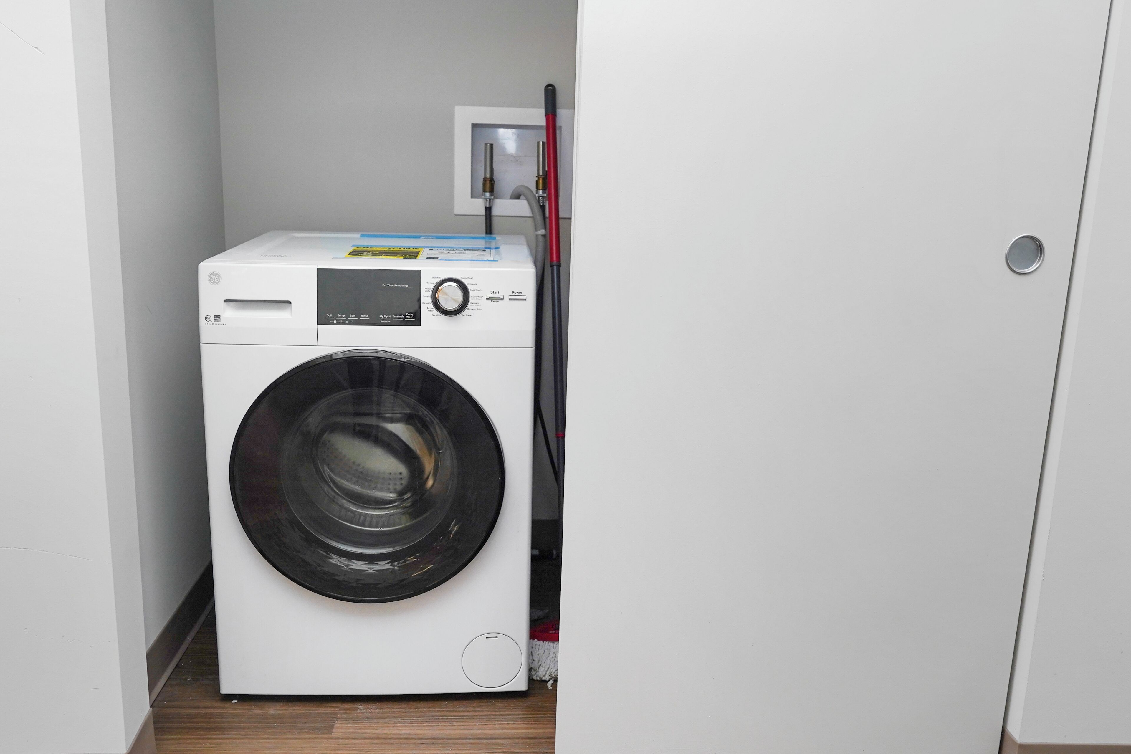 Una lavadora blanca y compacta en un lavadero pequeño, con una puerta a la derecha y un panel de control fijado a la pared.