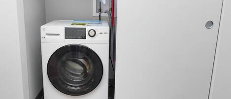 Una lavadora blanca y compacta en un lavadero pequeño, con una puerta a la derecha y un panel de control fijado a la pared.