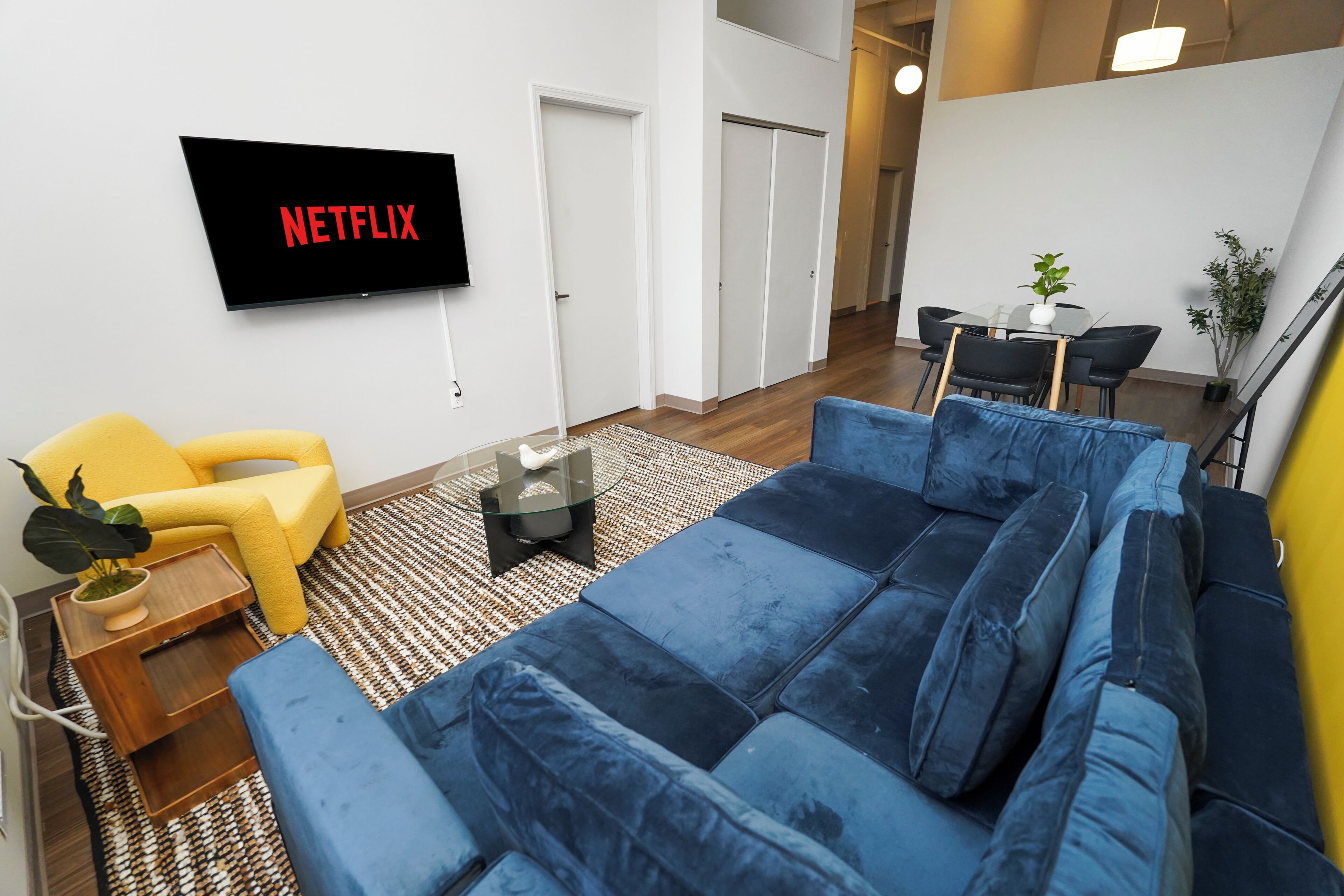 Una sala de estar con un sofá azul, un sillón amarillo, un televisor de pantalla plana mostrando 'NETFLIX', una mesita con una planta y un comedor al fondo.