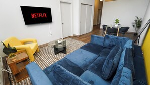 A living room with a blue sofa, a yellow armchair, a flat-screen TV displaying 'NETFLIX,' a small table with a plant, and a dining area in the background.