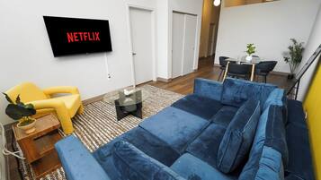 Una sala de estar con un sofá azul, un sillón amarillo, un televisor de pantalla plana mostrando 'NETFLIX', una mesita con una planta y un comedor al fondo.