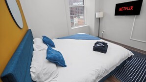 A hotel room with a bed, a TV displaying 'NETFLIX,' a window with a view of buildings, and a lamp.