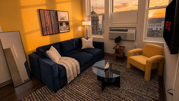 A living room with a sofa, armchair, coffee table, and a TV. There are windows with a cityscape view, a wall-mounted air conditioner, and a lamp.