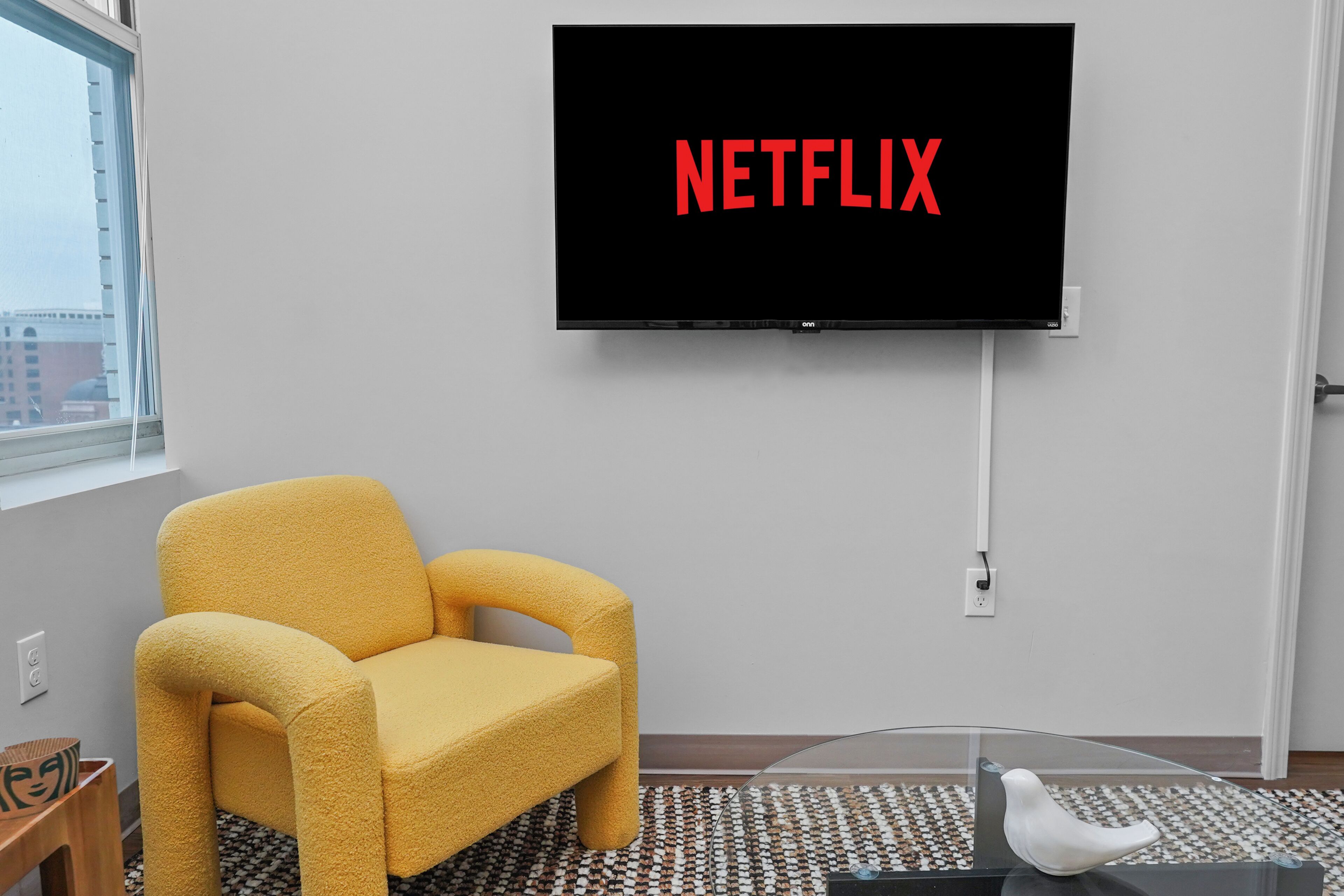 Una habitación con una televisión de pantalla plana grande montada en la pared que muestra 'NETFLIX', un sillón amarillo, una mesa de centro de vidrio y una alfombra estampada.