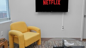 A room with a large flat-screen TV mounted on the wall displaying 'NETFLIX,' a yellow armchair, a glass coffee table, and a patterned rug.