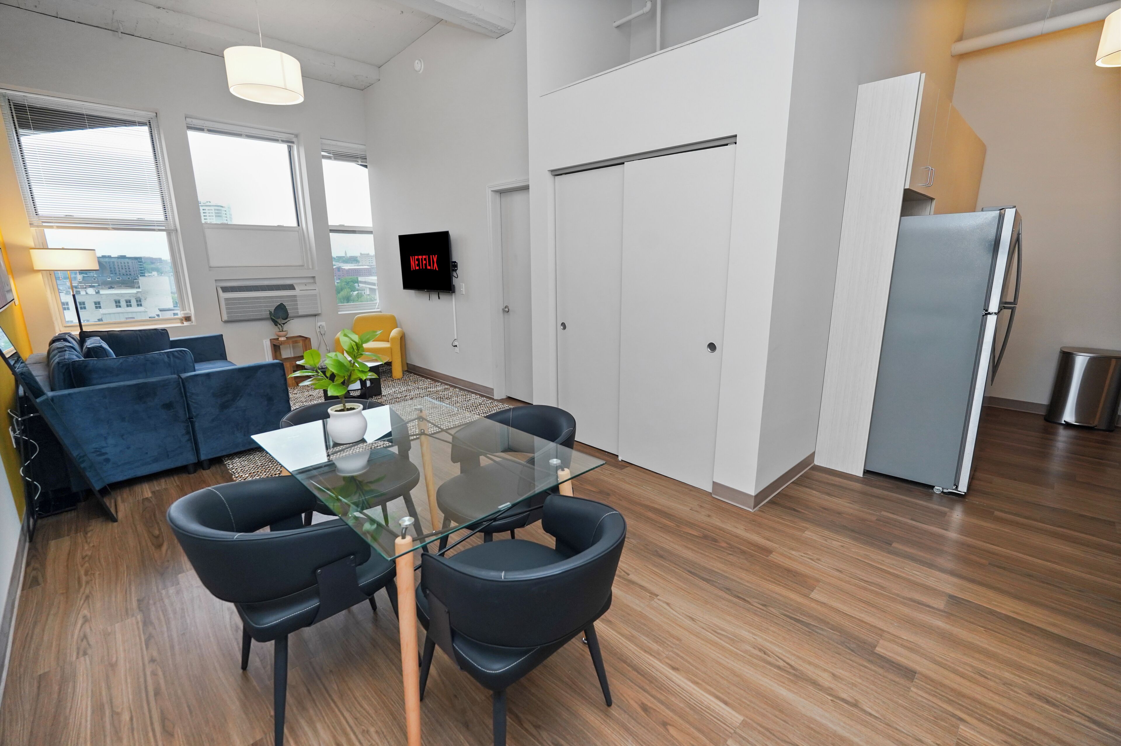 Un espacio habitable moderno con una mesa de comedor de vidrio, sillas negras, un sofá azul y un refrigerador blanco.