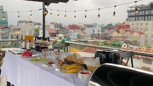 A rooftop dining area with a table set for breakfast, a grill, and a cityscape view.