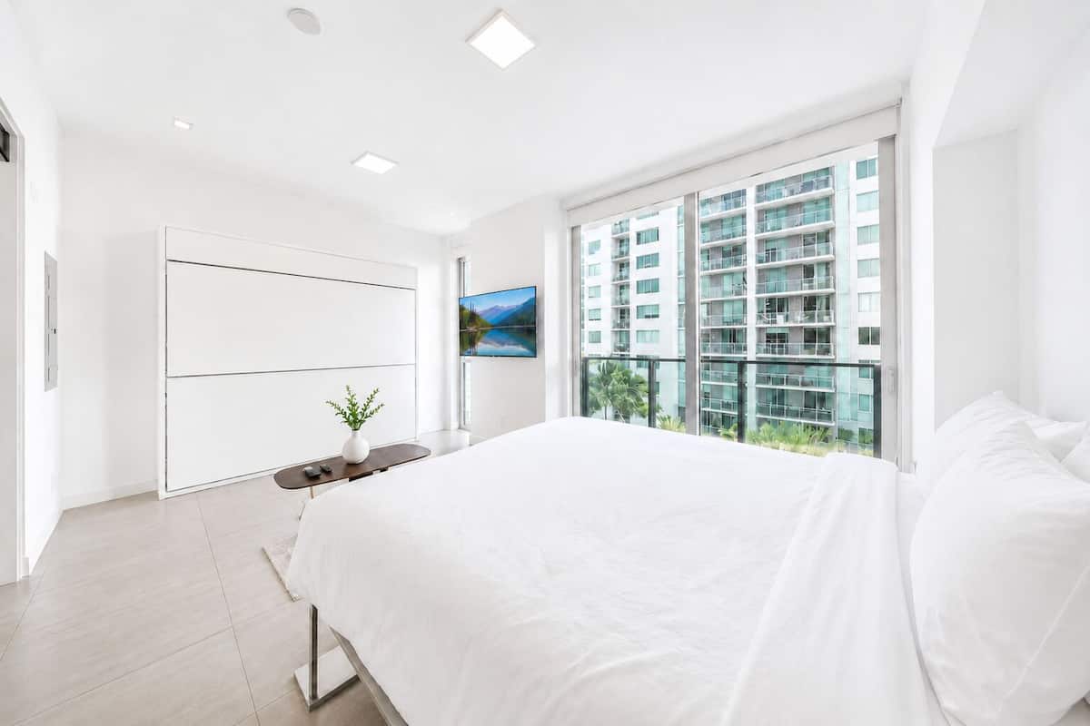 Quarto moderno com uma cama grande, televisão e varanda com vista para a cidade.