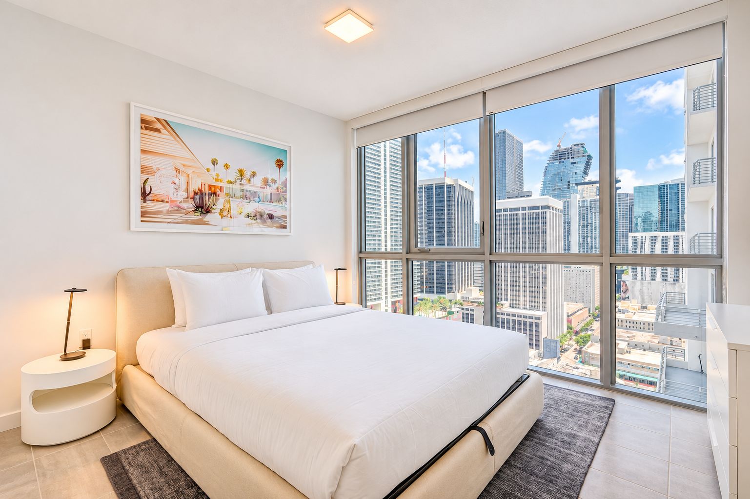 A modern hotel room with a large bed, a city view, and a framed artwork on the wall.
