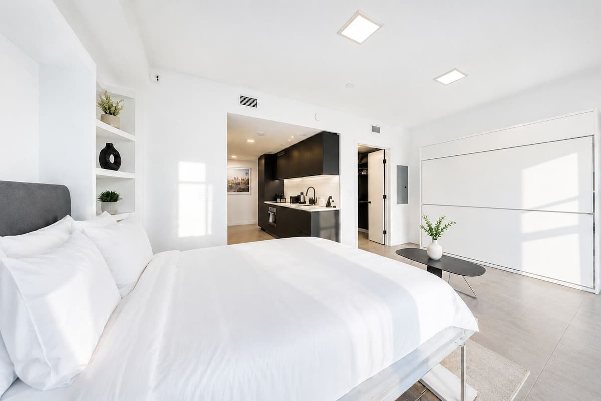 Quarto moderno com cama grande, secretária embutida e vista para uma cozinha.