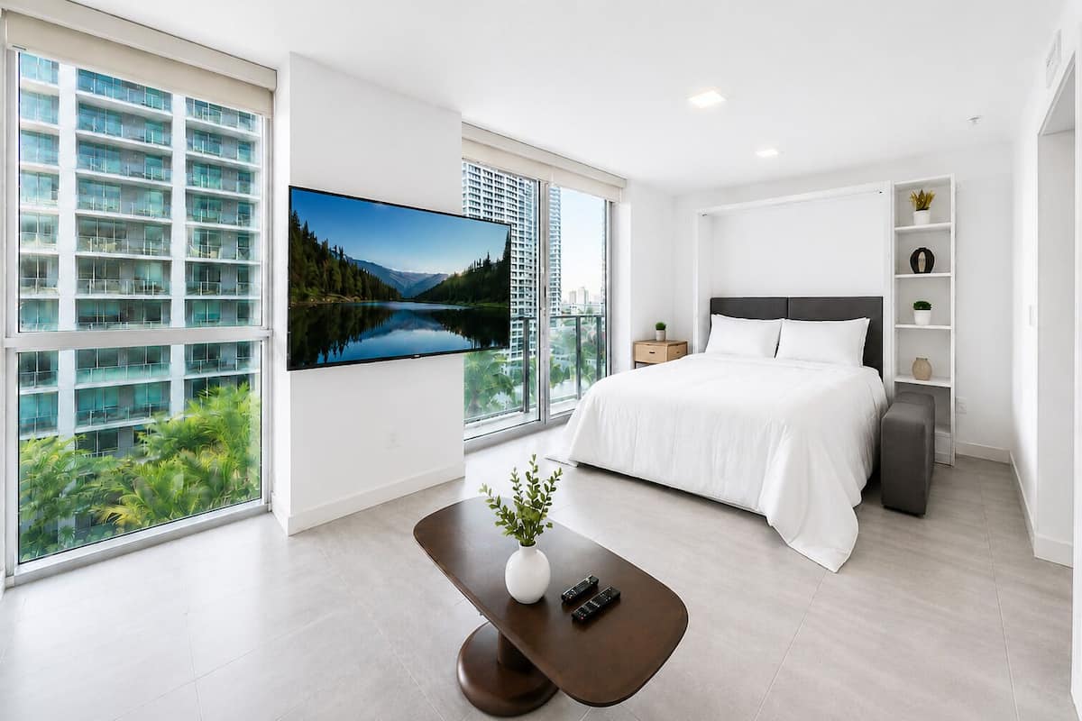 Quarto moderno com uma cama grande, televisão na parede, uma mesinha com uma planta e vista para um edifício e áreas verdes.