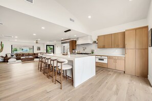 A spacious kitchen with wooden cabinets, a central island with stools, and a large window providing a view of the outdoors. - Luxury Pool Oasis - Modern Desert Home w/ Resort Backyard, Fire Pits & Game Room (Scottsdale)