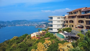 A coastal hotel with multiple floors, a swimming pool, and a view of the sea and cityscape.