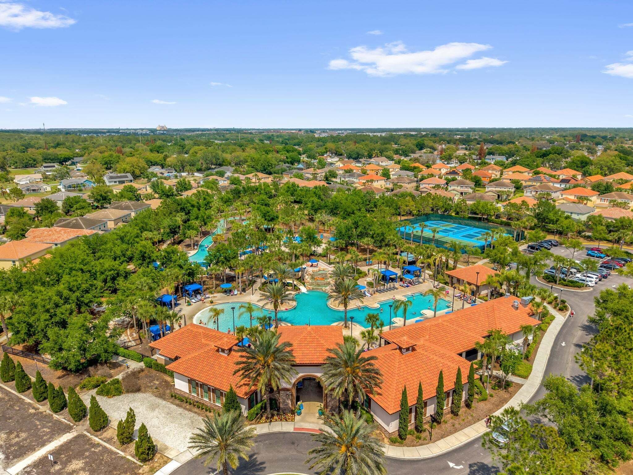 A residential area with a large swimming pool surrounded by houses with red-tiled roofs and palm trees.