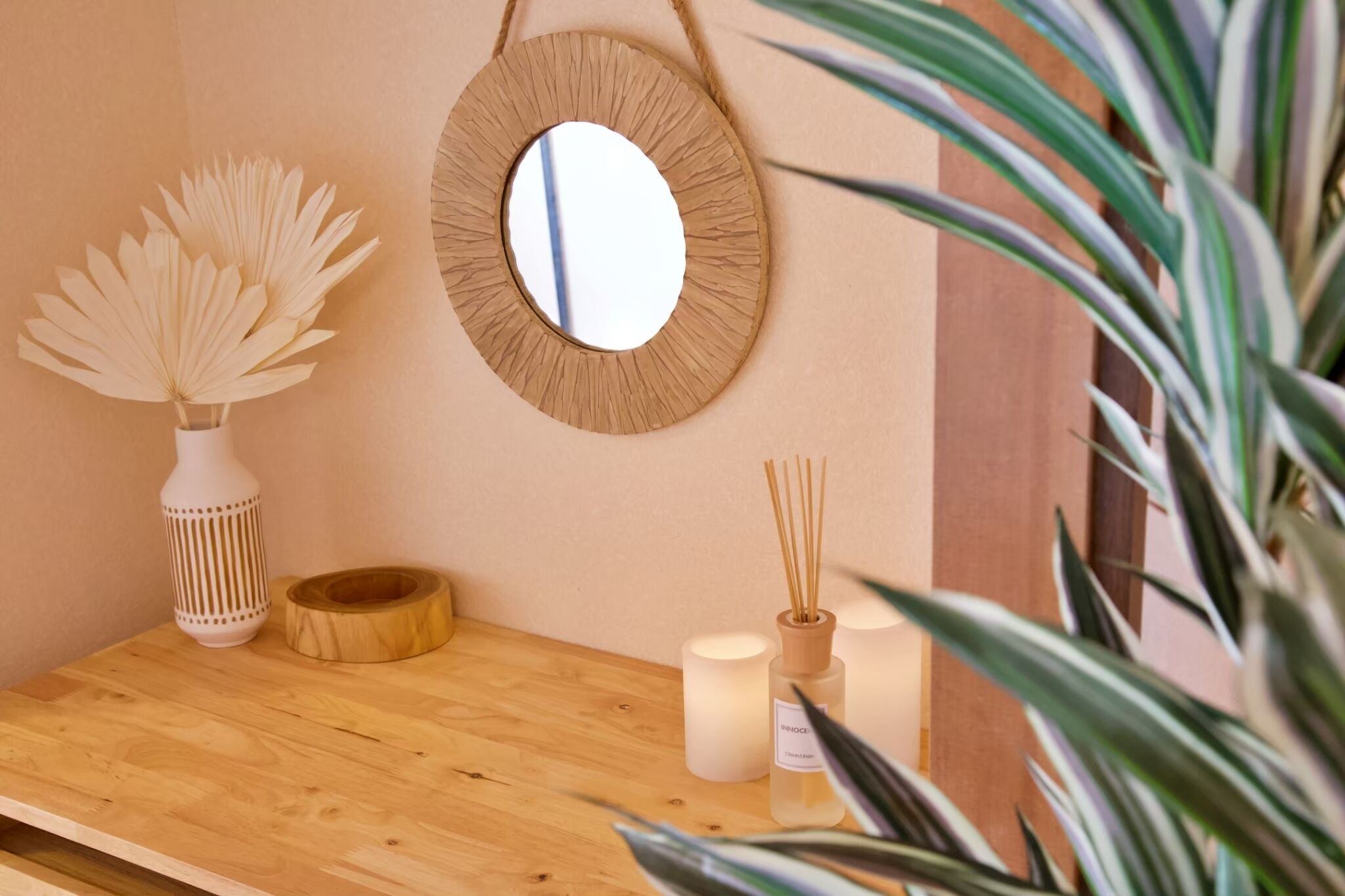 A wooden shelf with a mirror, a vase with dried flowers, candles, and reed diffusers.