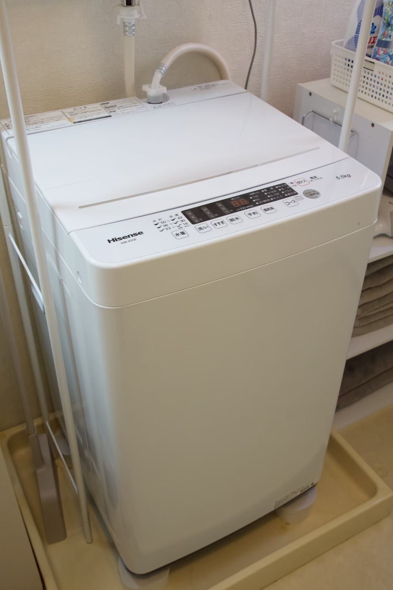 A white Hisense washing machine with a control panel, placed in a room with a white wall and a white shelf.