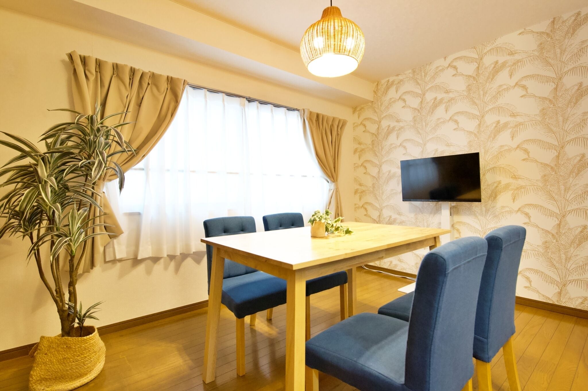 Ein Zimmer mit einem Holzesstisch, blauen Stühlen, einer Topfpflanze, einem Fernseher an der Wand und einer Wandtapete mit Muster.