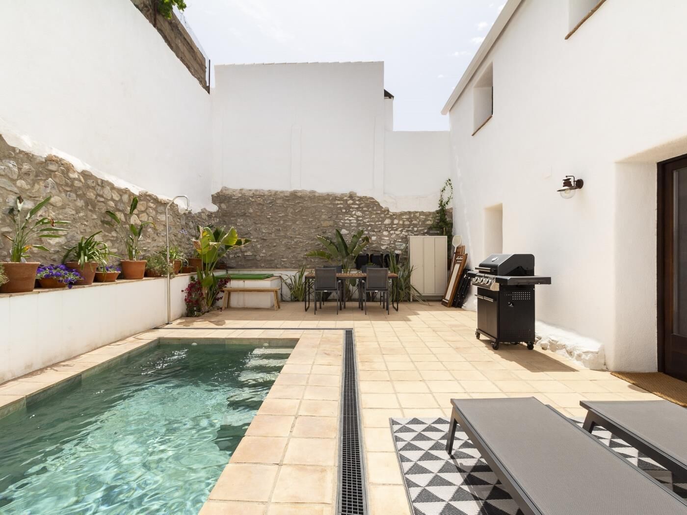 A pool area with a stone wall, potted plants, a barbecue, and outdoor furniture.