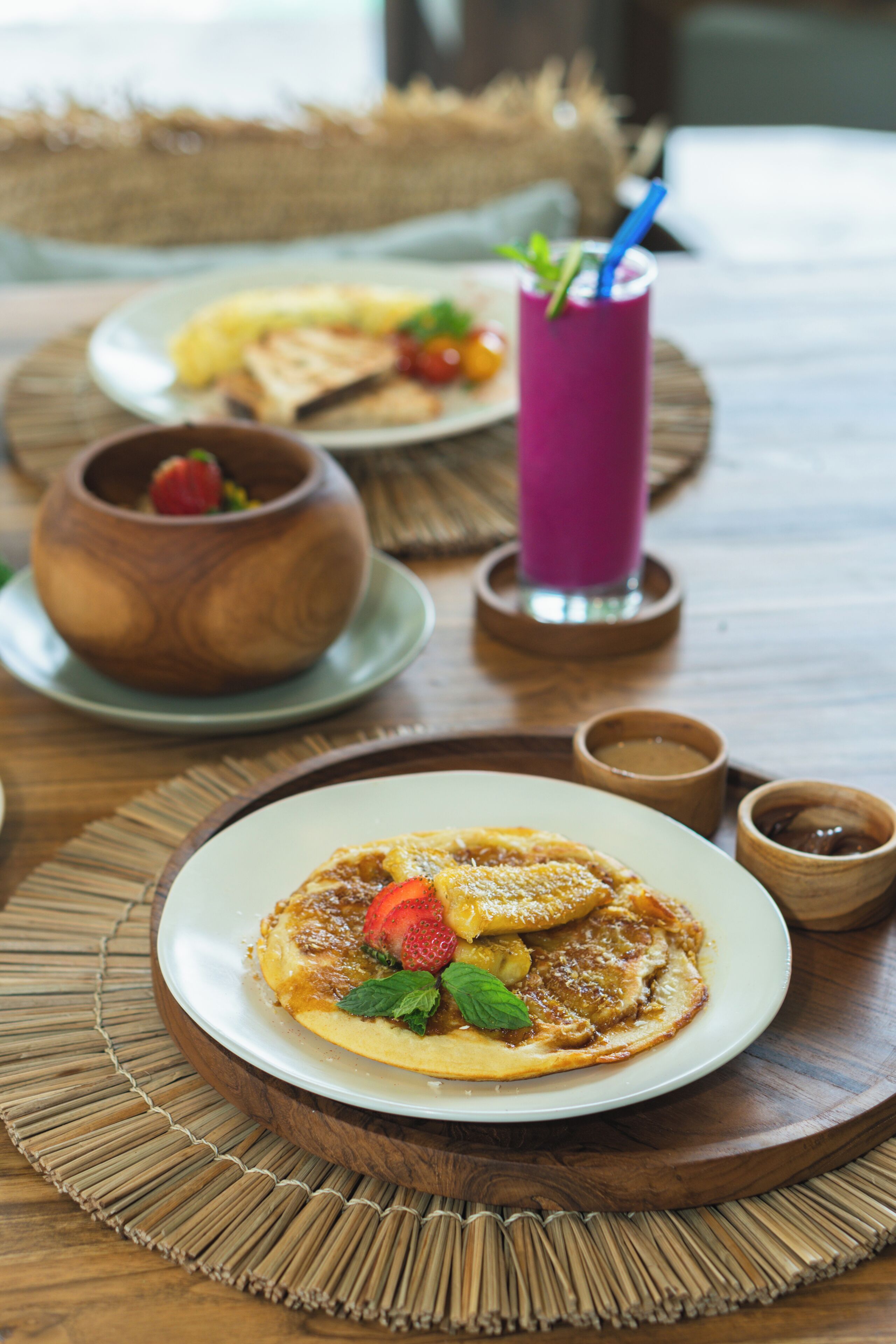 A dining table with a plate of pancakes topped with strawberries and mint, a purple drink, and a small wooden bowl.
