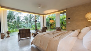 Un dormitorio espacioso con una cama grande, una silla de madera, una mesita y vistas a un follaje exuberante.