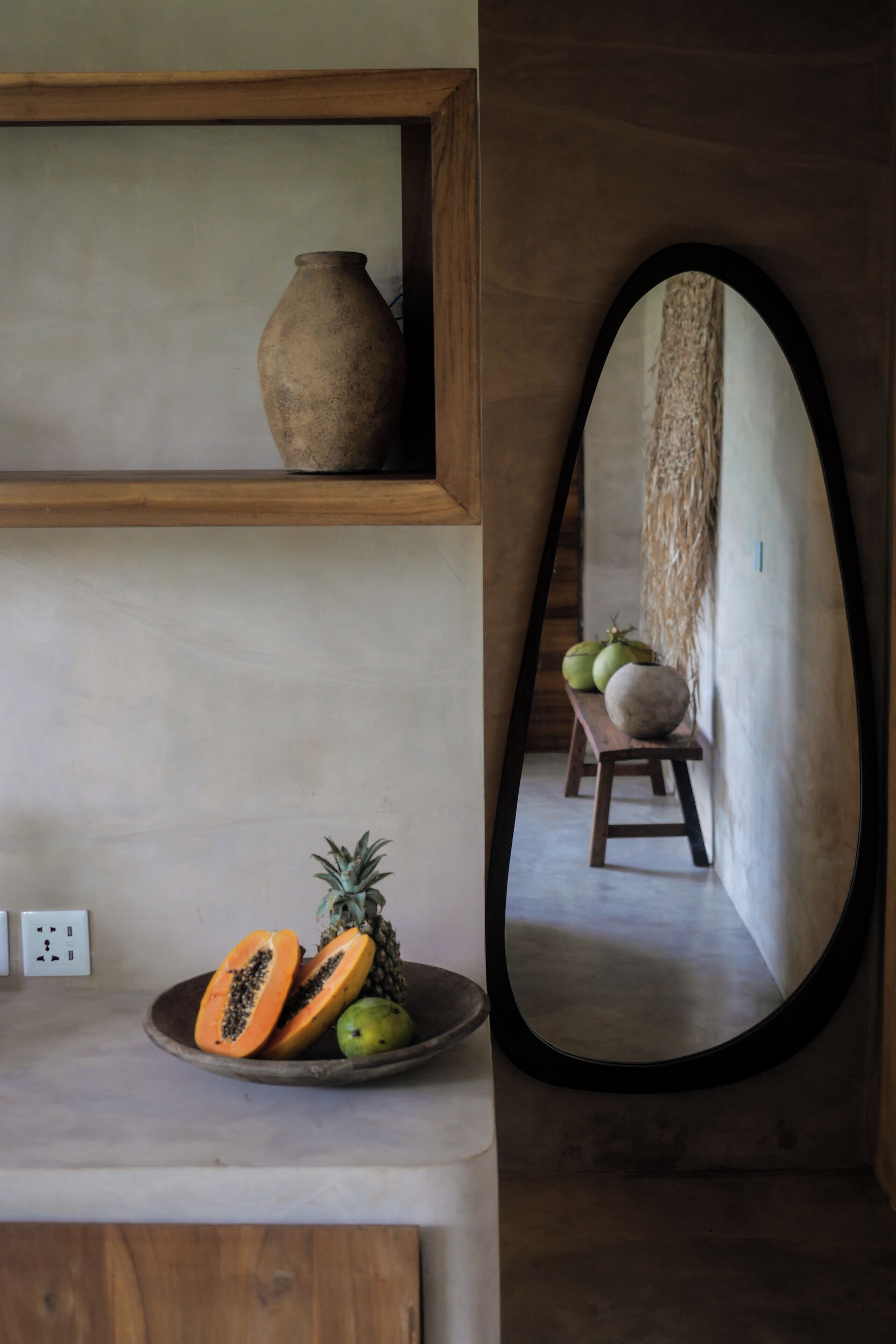 A kitchen counter with a bowl of fruit, a wooden shelf with a vase, and a mirror reflecting a room with a bench and wall decor.