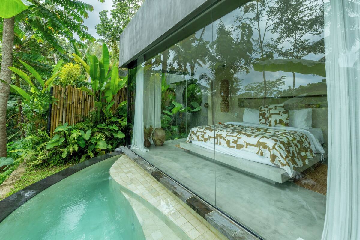 Un dormitorio con cama, pared de vidrio y vista a un jardín.
