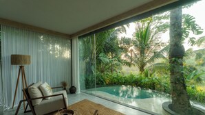 Una habitación moderna con pared de vidrio, sofá, lámpara y vista a una piscina rodeada de plantas tropicales.