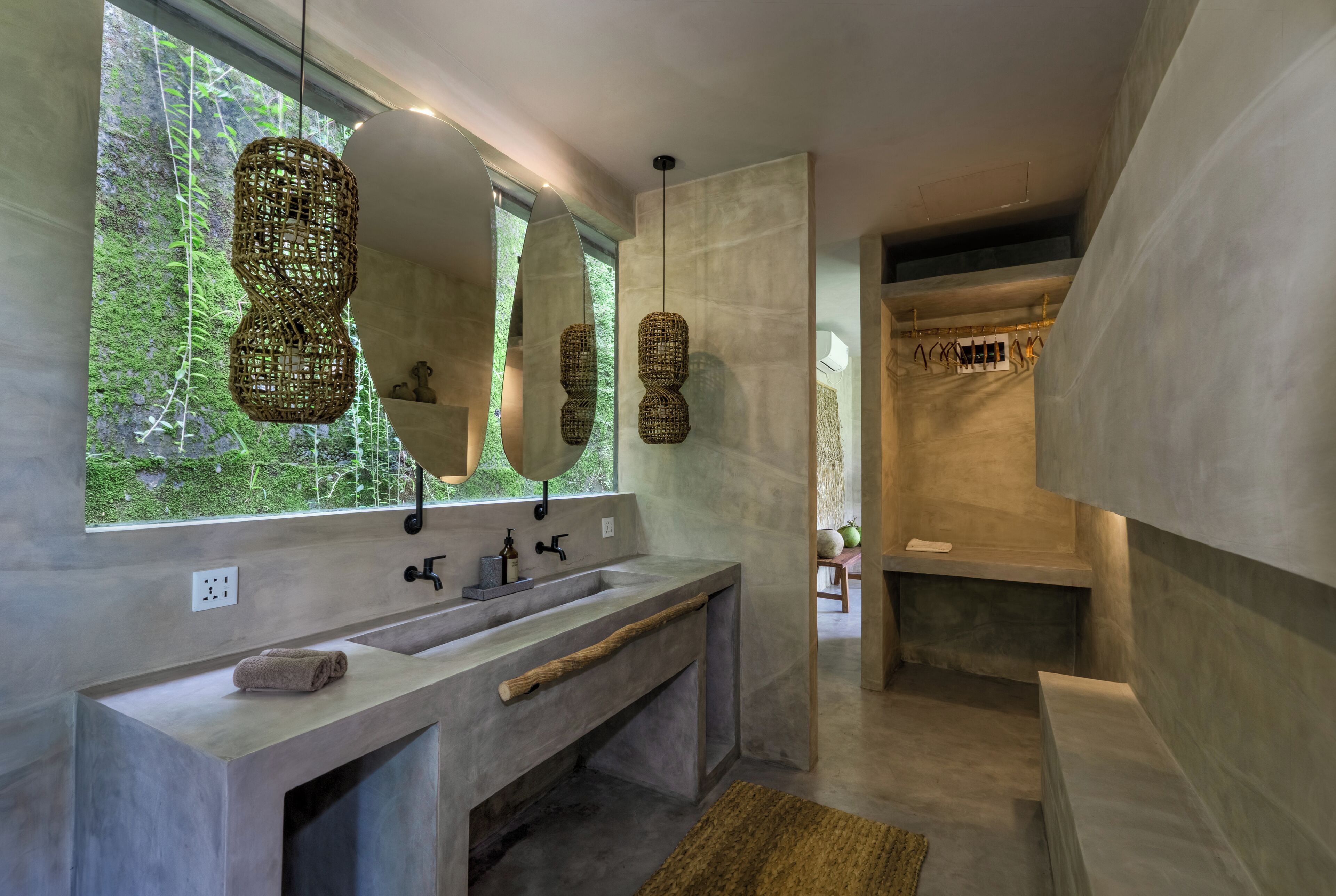 A modern bathroom with a large mirror, two sinks, a wooden bench, and a view of greenery outside.