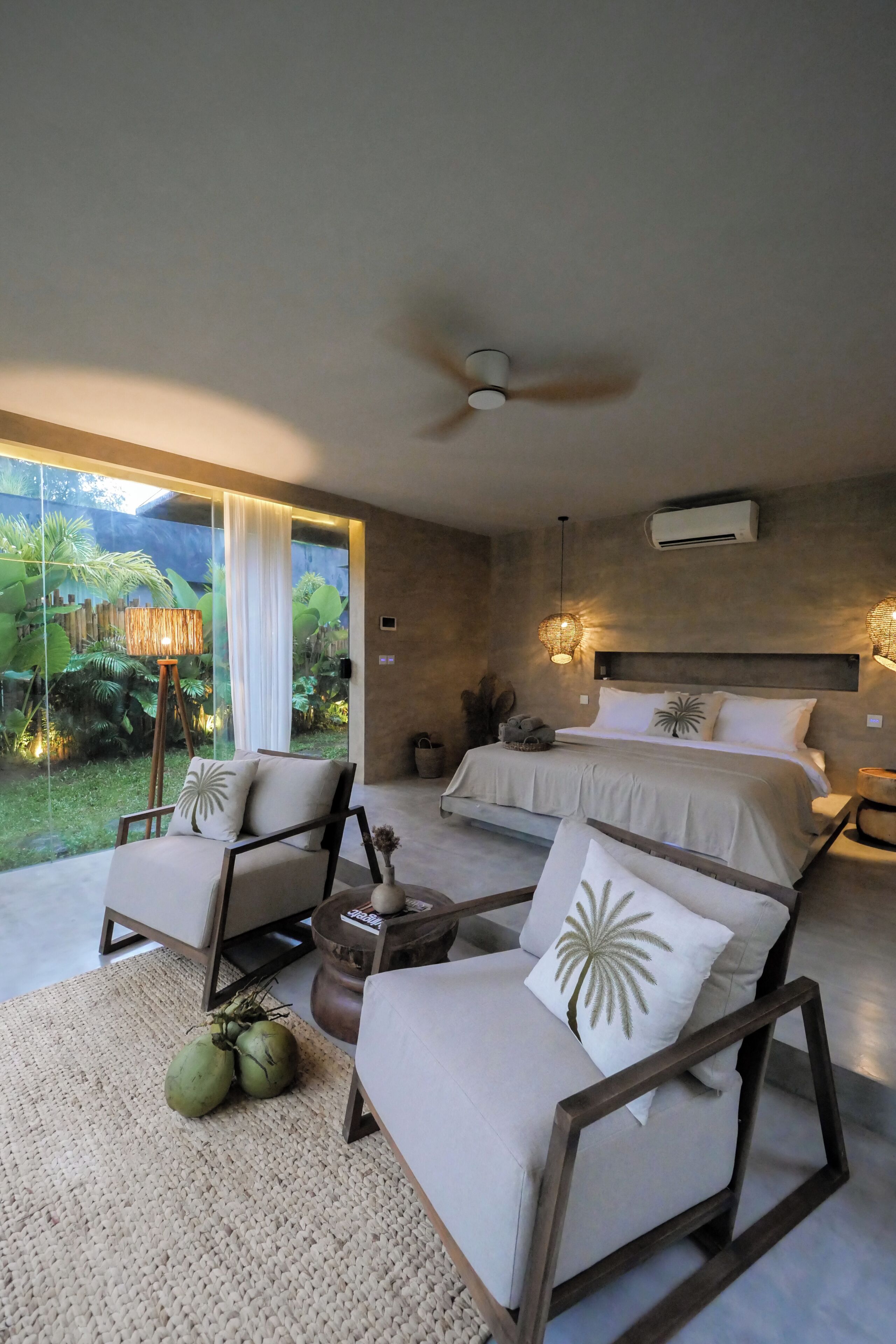 A bedroom with a bed, two chairs, a ceiling fan, and a view of greenery.