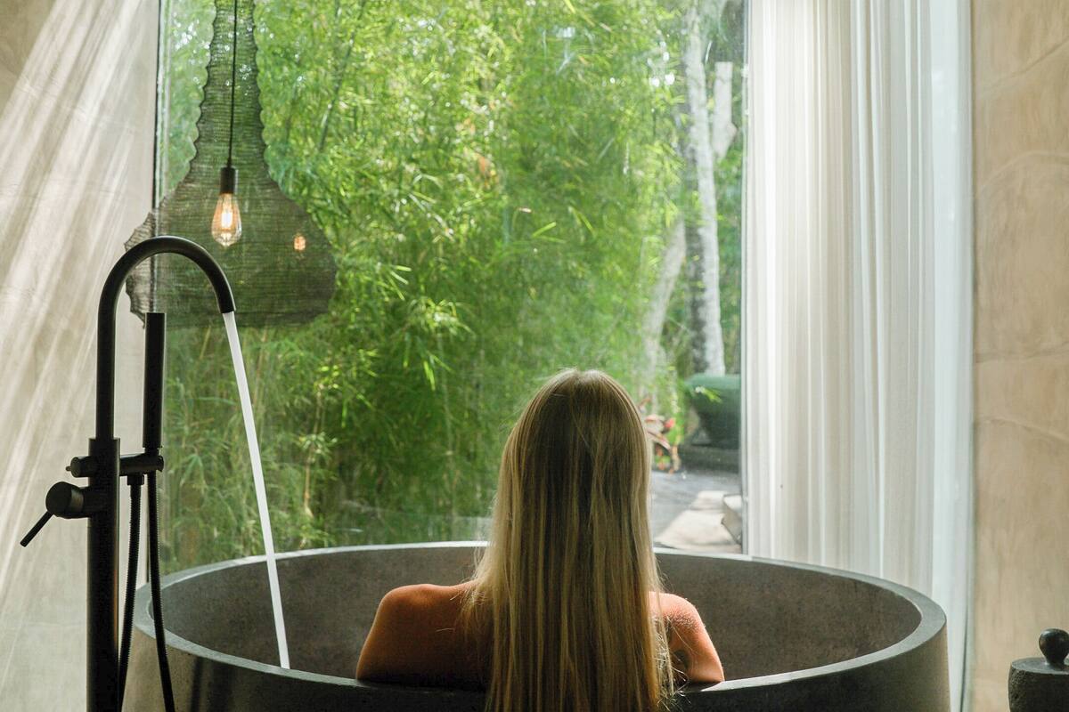 Una persona sentada en una bañera grande y redonda, con vistas a la vegetación exterior.