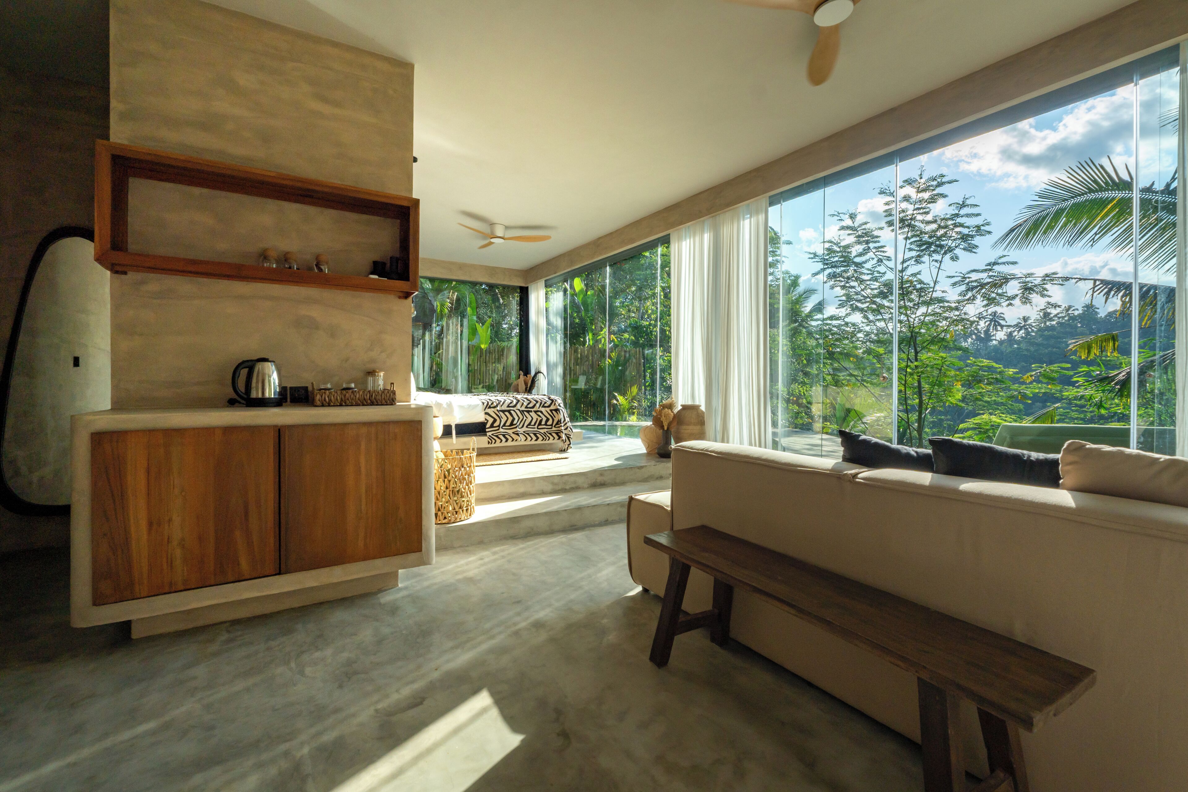 Una sala moderna con sofá, banco de madera, zona de cocina con cafetera y ventanas amplias que ofrecen vistas de abundante vegetación.