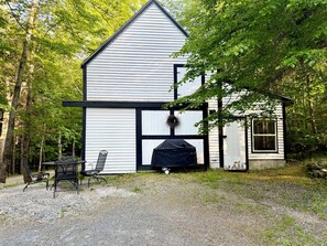 Un bâtiment blanc avec des bordures noires, un espace barbecue couvert et une table avec des chaises à l’extérieur.
