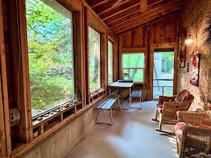 A rustic cabin interior with large windows, wooden beams, a picnic table, wicker furniture, and a stone wall.