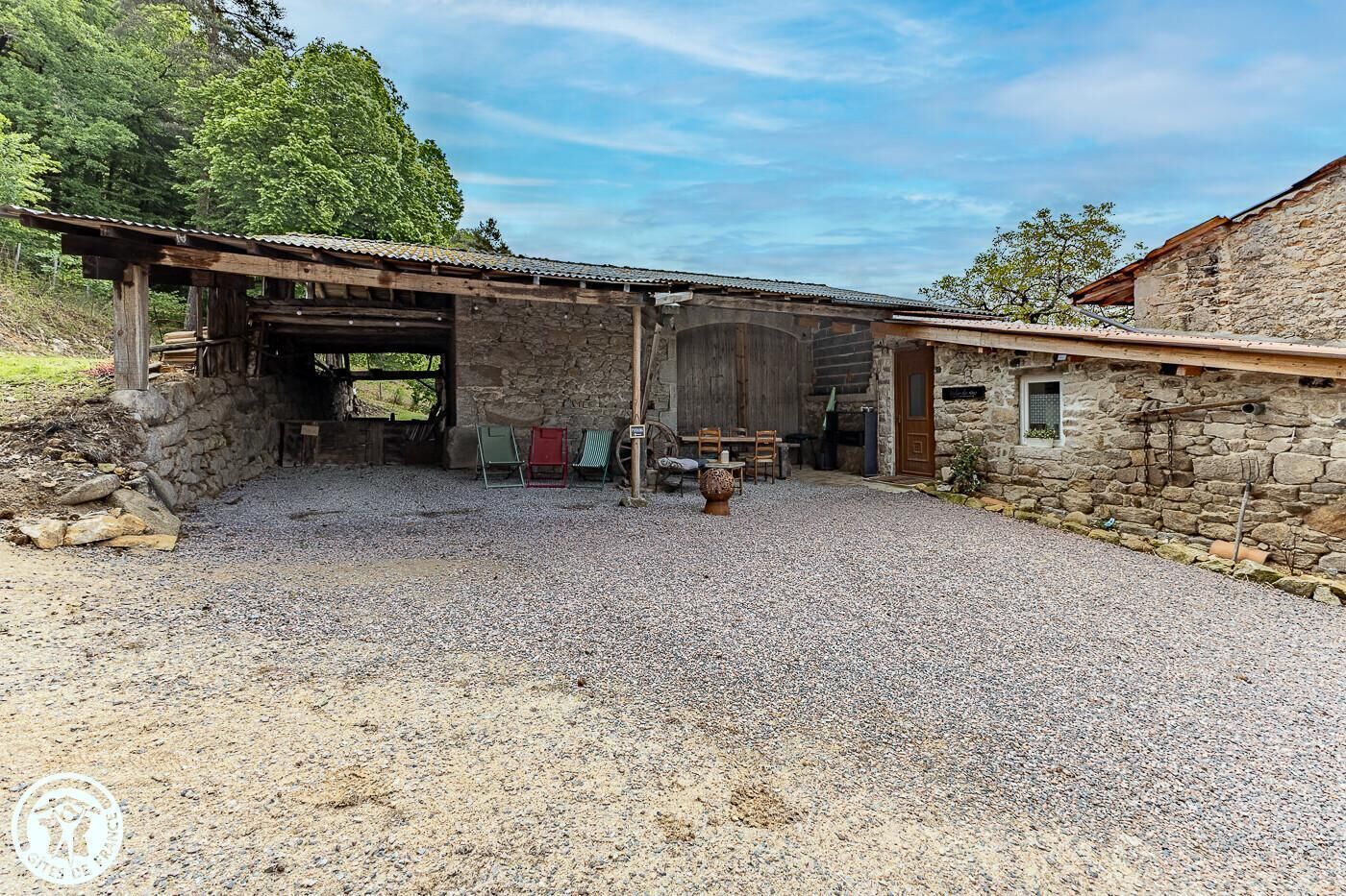 Un bâtiment rustique en pierre, avec une allée de gravier, des bancs en bois et un espace abrité comprenant une table et des chaises.